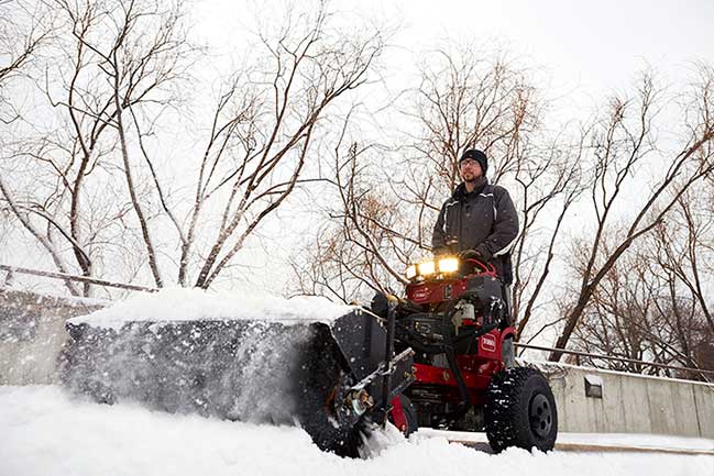 Toro Multi-Force stand-on machine with power broom clearing a snowy walkway